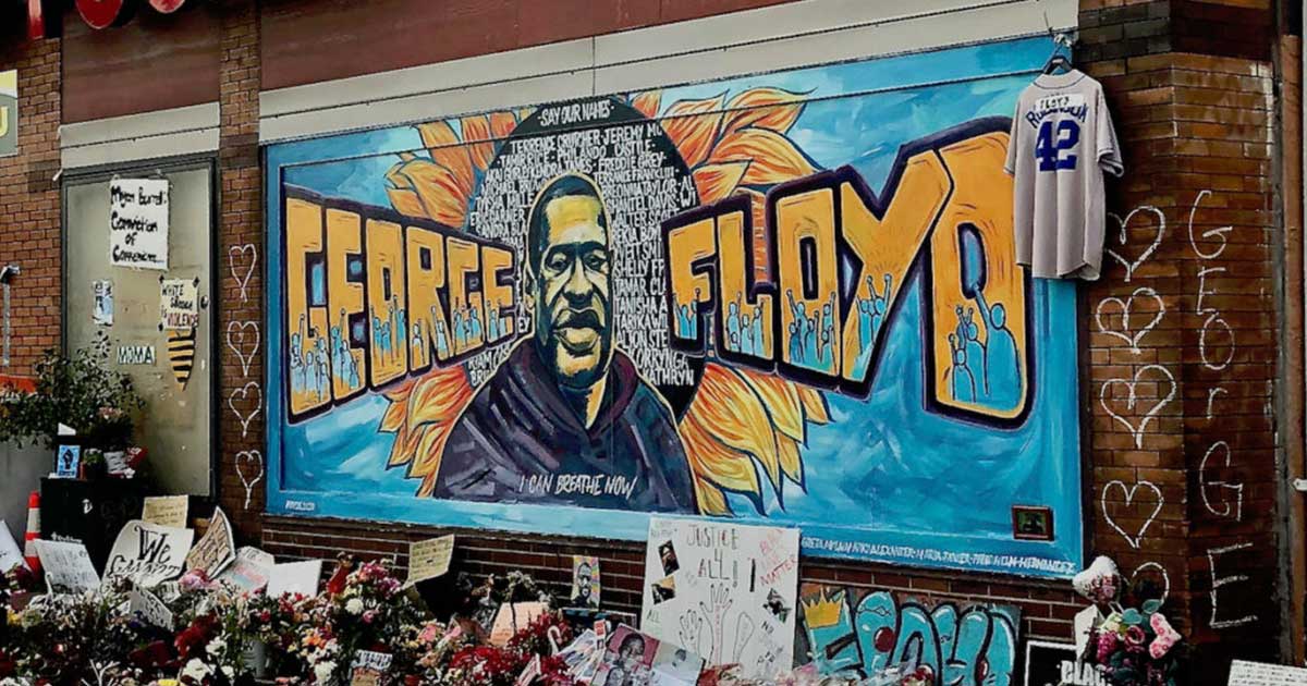 brightly-colored mural on a building with the name and face of George Floyd, surrounded by flowers and protest signs
