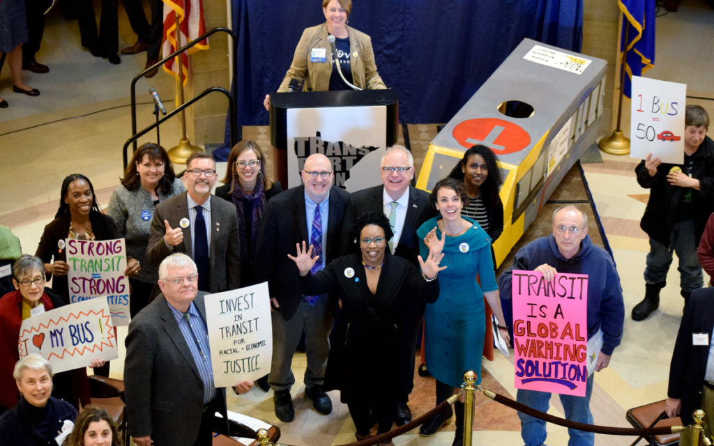 Governor Walz posing with legislators and transit advocates with pro-transit signs, at the Capitol Rotunda