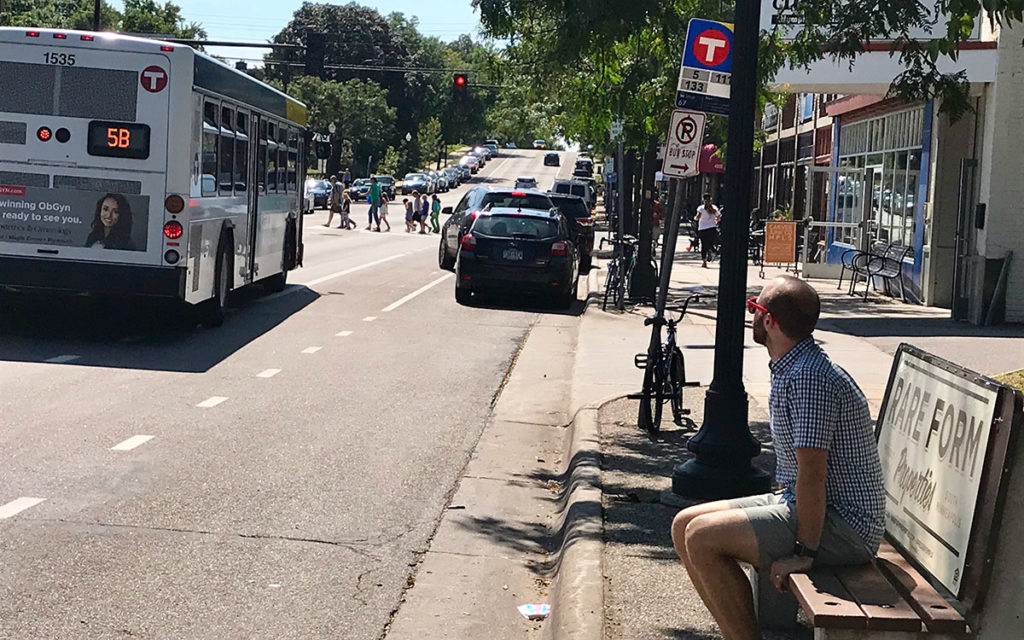 Minneapolis resident sits at southbound bus stop at 48th Street and Chicago Avenue as Metro Transit Route 5 bus passes by. Bus stop is a sign and bench only, no shelter. In the background there are businesses and people walking across the street.