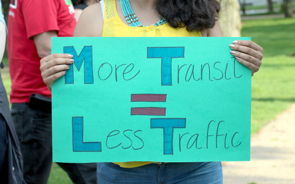 Woman at a climate rally holds a sign that says "More Transit = Less Traffic"