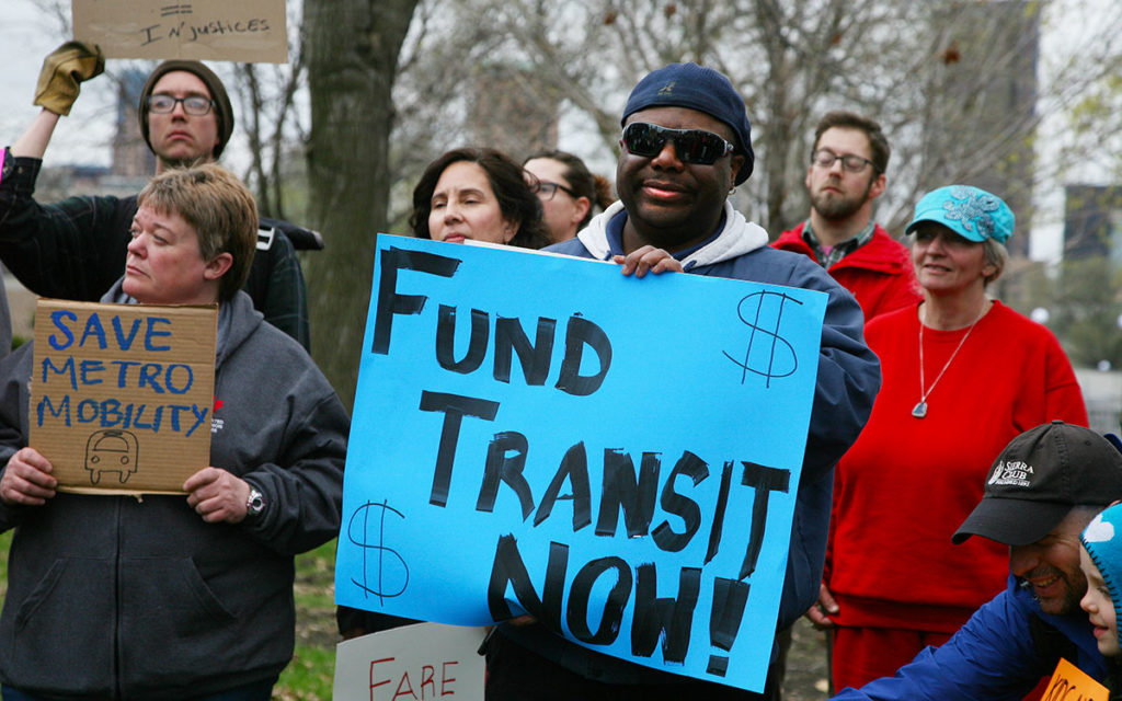 Transportation options advocates outside the MN State Capitol. Man holds sign that says "Fund Transit Now!" Woman holds sign that says "Save Metro Mobility."