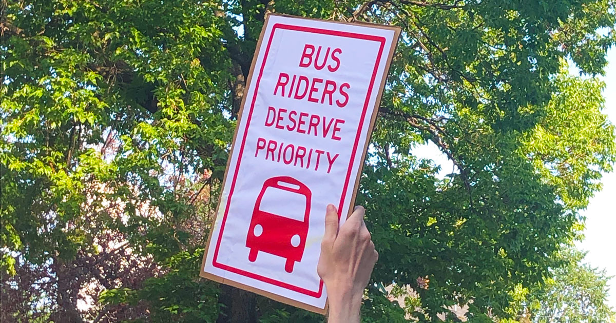 A person's hand holds up a rally sign that reads Bus Riders Deserve Priority.