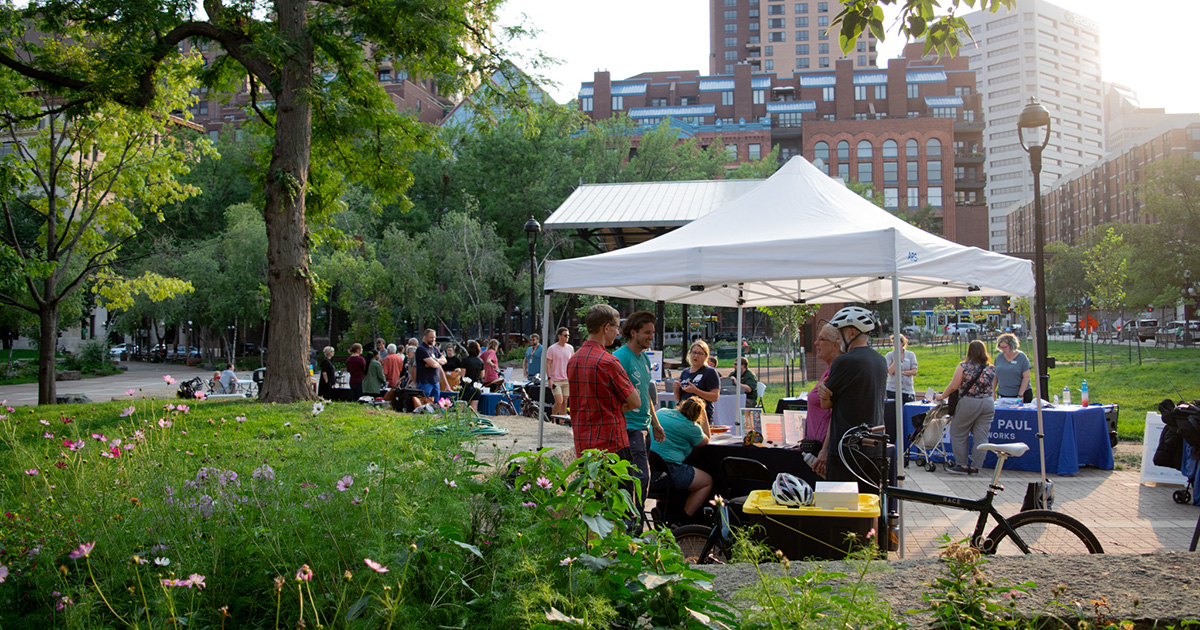 Move Minnesota connects with community members at the Saint Paul Mobility Mingle in Mears Park. Flowers, trees, and tall buildings frame the image.