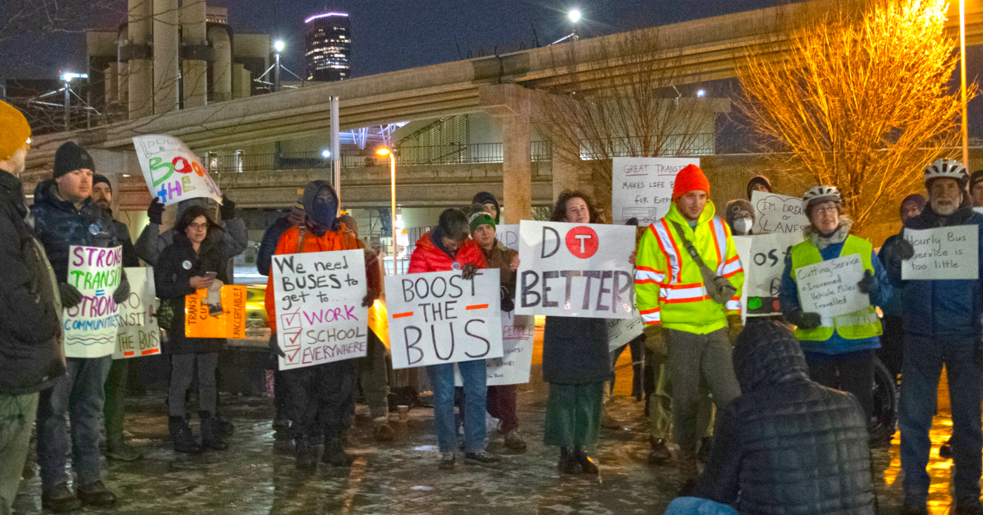 A group of people rally in the cold with colorful signs reading "Boost the Bus" and "We need buses to get to work, school, everywhere!"