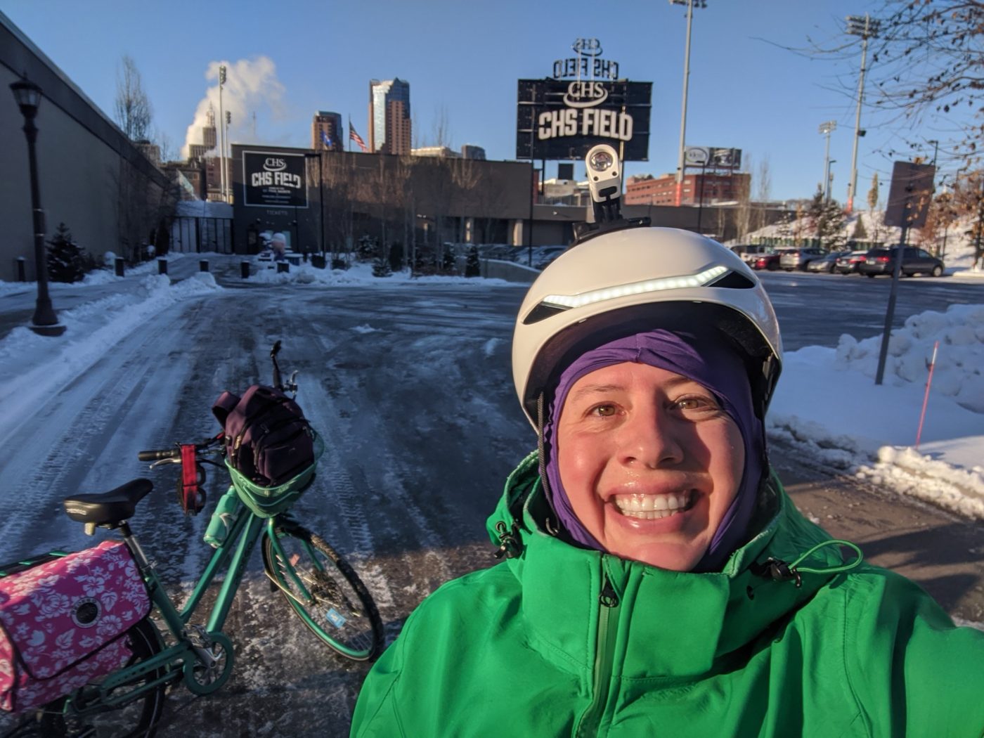 A woman in a bike helmet smiles with their bicycle while stopped on a winter day in Saint Paul, Minnesota. CHS Field is behind them. Snow is on the ground all around.