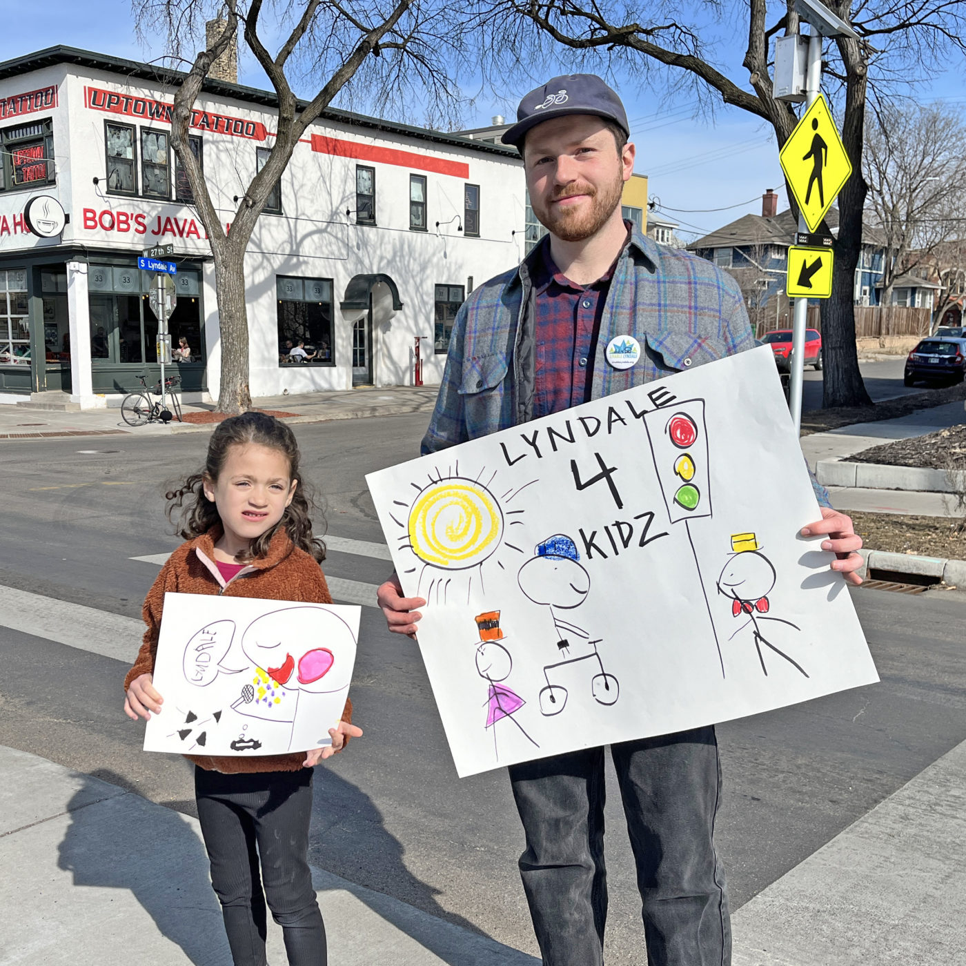 Phil Schwartz, a white man with a brown beard, stands next to his young daughter, Holly, with a sign that says "Lyndale 4 Kidz" over a drawing of stick figure people walking and riding bikes under a yellow sun