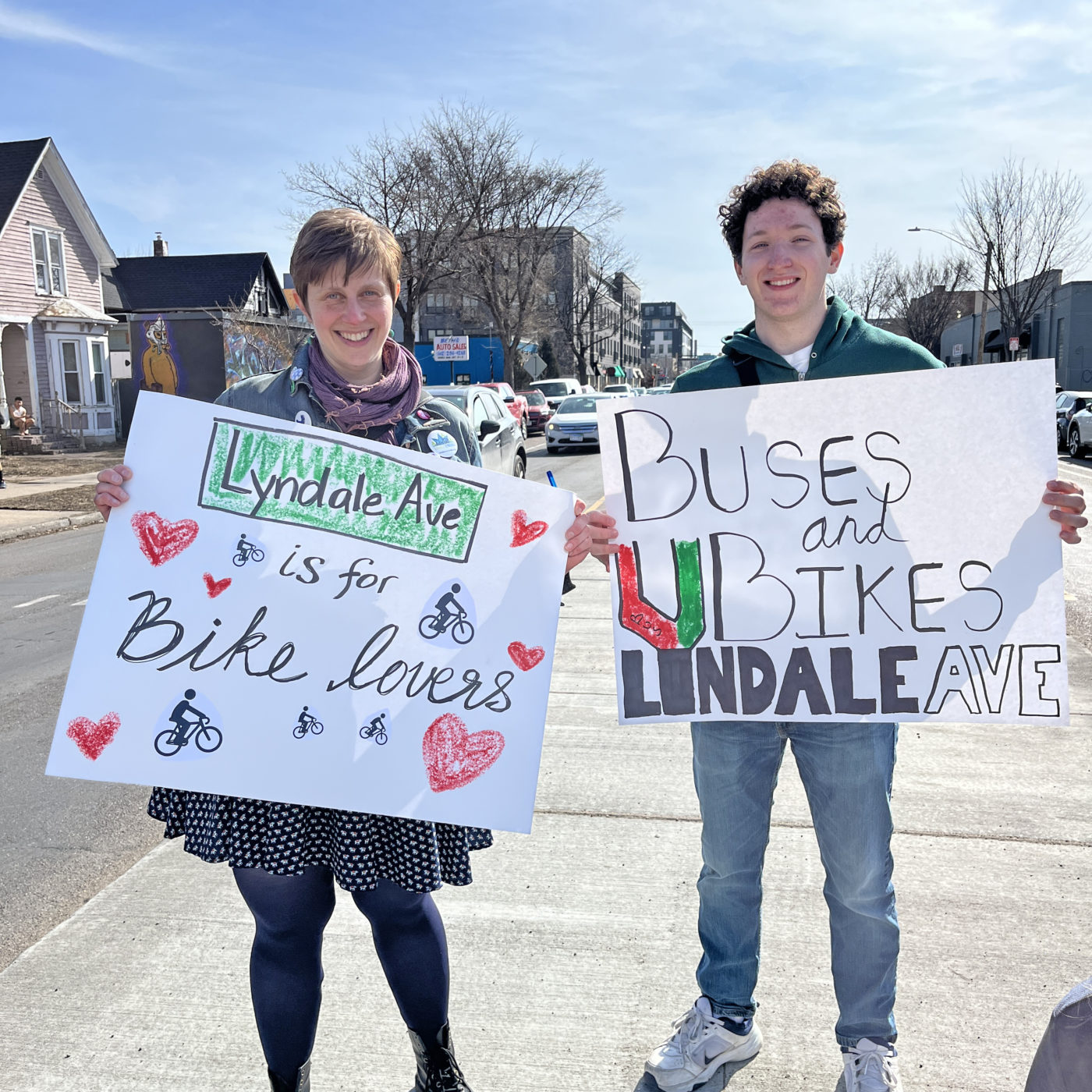 Two community members, including Asa Stanfield, hold signs in the median at 27th and Lyndale that say Lyndale Ave is for Bike Lovers" and "Buses and Bikes on Lyndale Avenue"