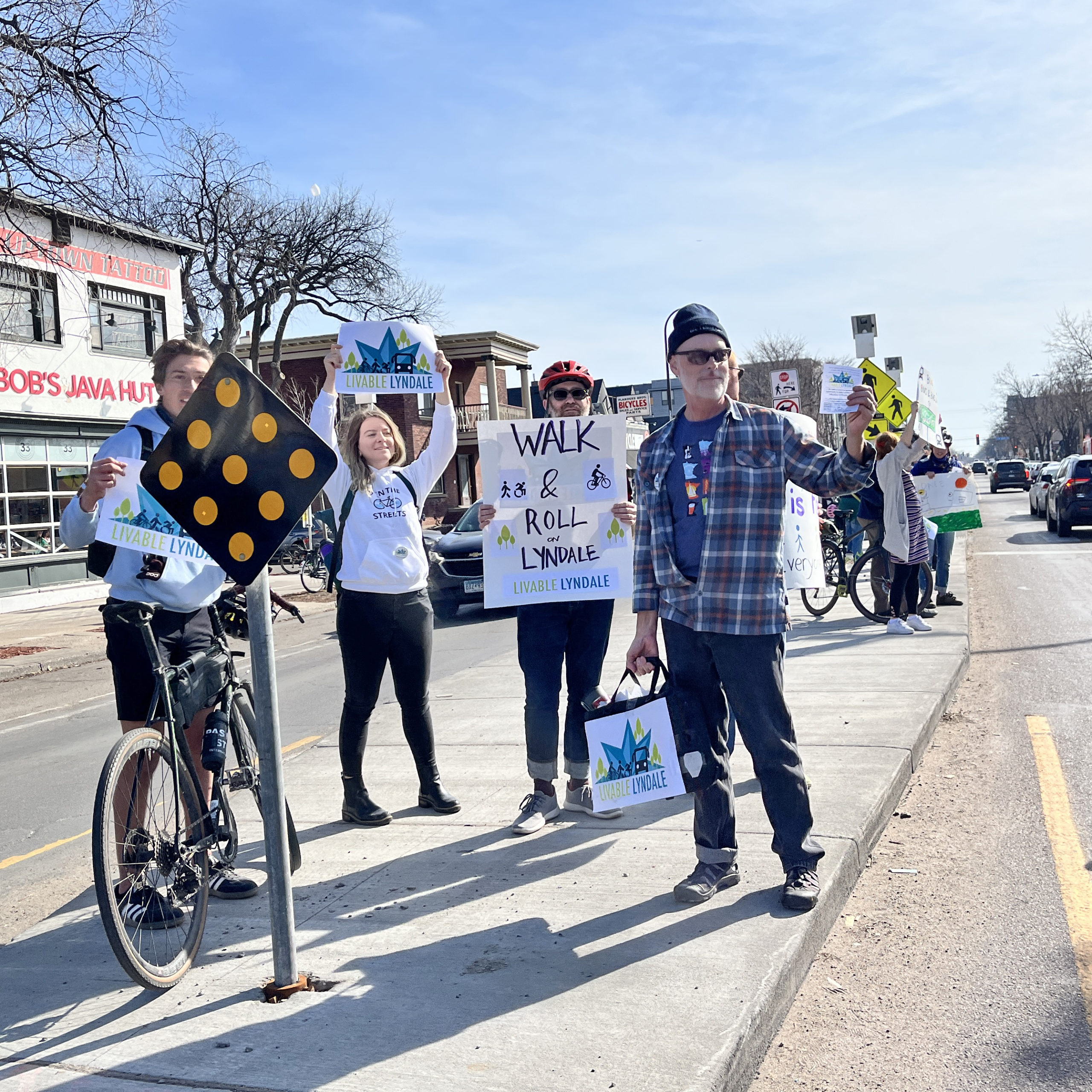 A group of community members hold Livable Lyndale signs while standing in the median together at Lyndale and 27th in Minneapolis