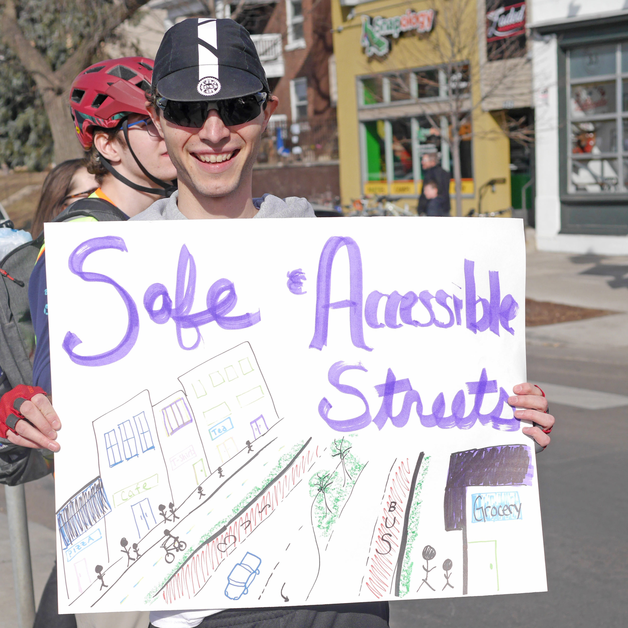 A community member wearing a black cycling cap and sunglasses holds a sign that says Safe and Accessible Streets over a drawing of a street with people walking, and biking next to businesses 