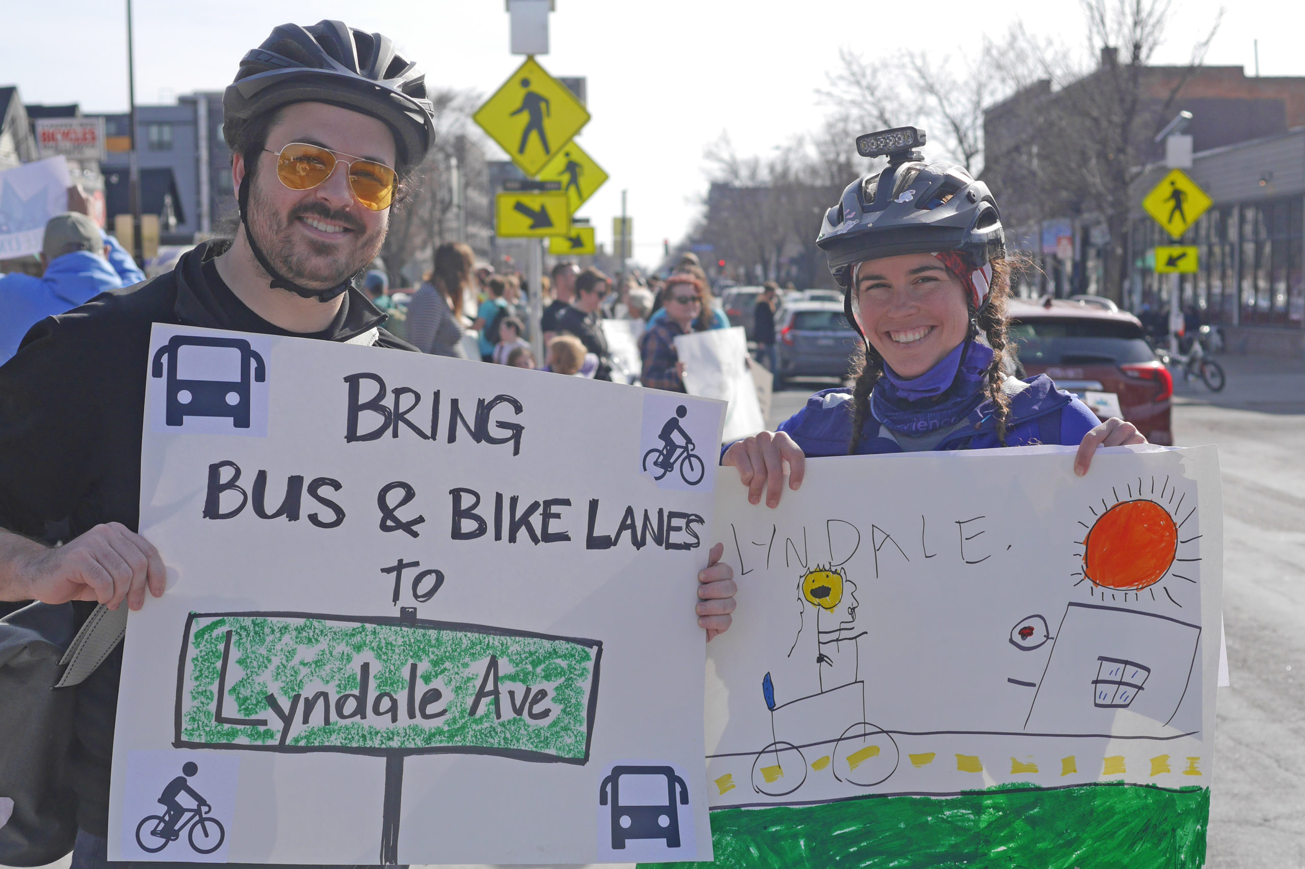 Community members in bike helmets at the Livable Lyndale rally in Minneapolis. Their signs say "Bring bus and bike lanes to Lyndale Ave" and a drawing of biking in a bike lane. A crowd of people at the rally is visible in the background.