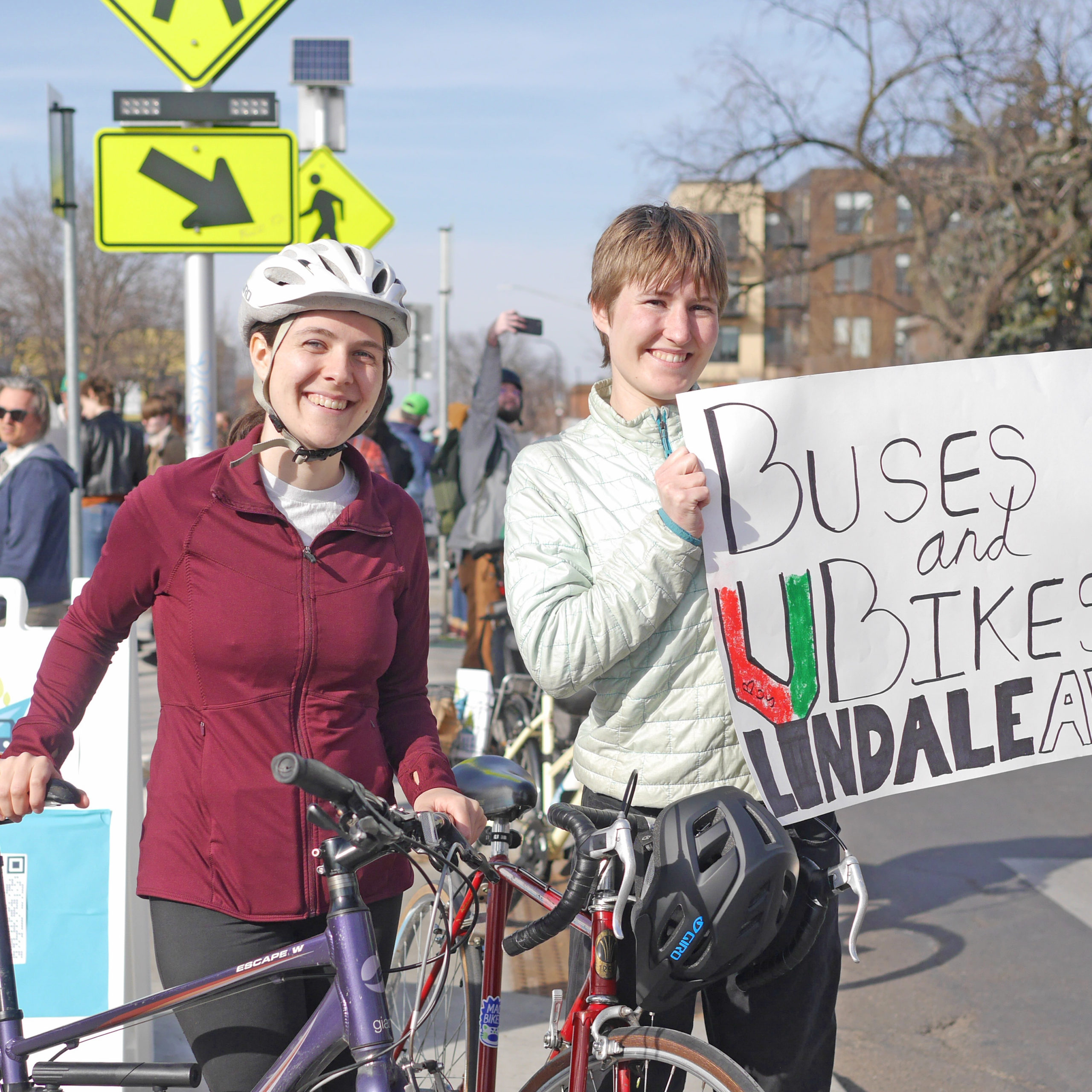 Two community members in the median at 27th and Lyndale, one standing with her purple bicycle and the other holding a sign that says Buses and Bikes on Lyndale Ave!