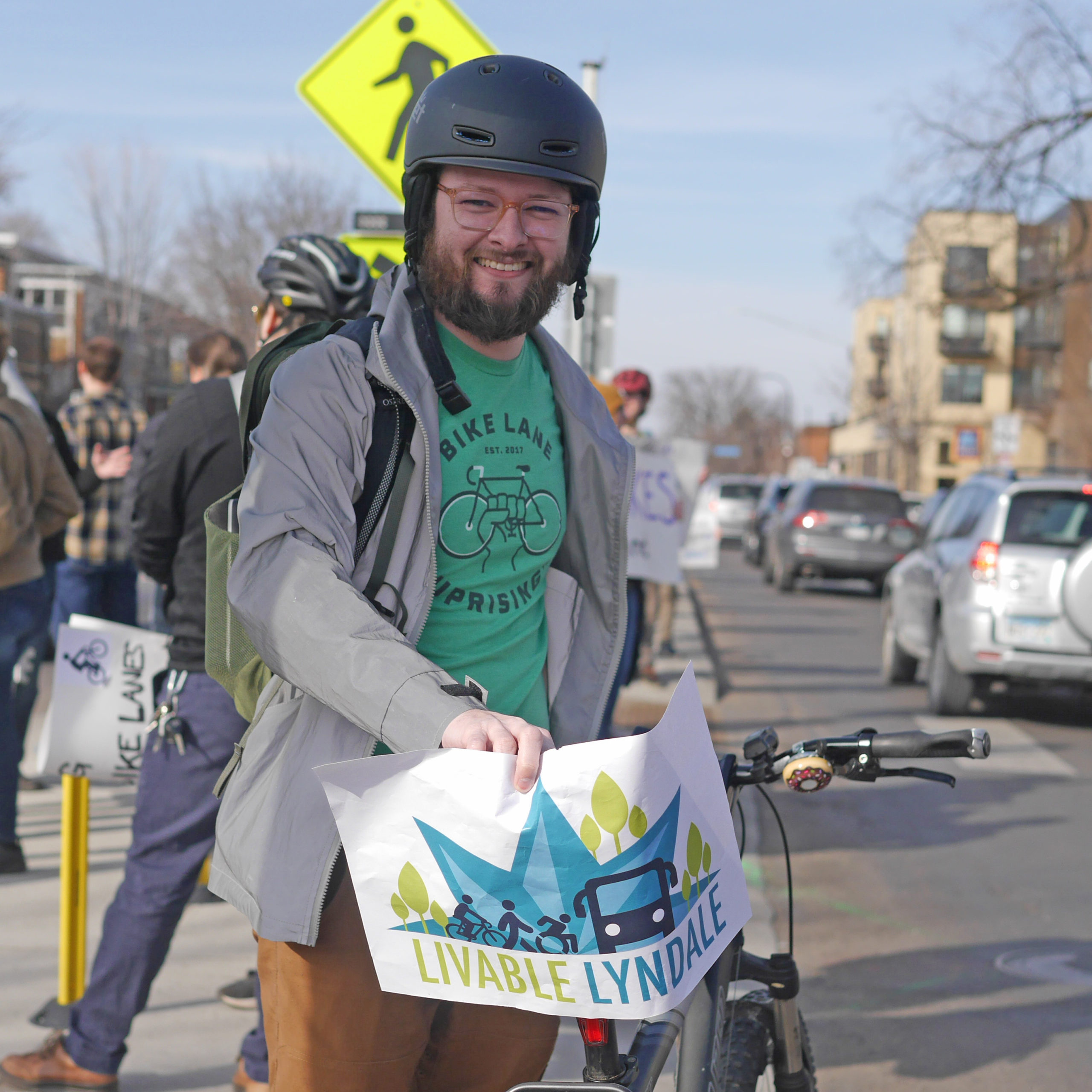 A community member wearing a black helmet and holding a Livable Lyndale sign on the back of his bike at the rally at 27th and Lyndale 