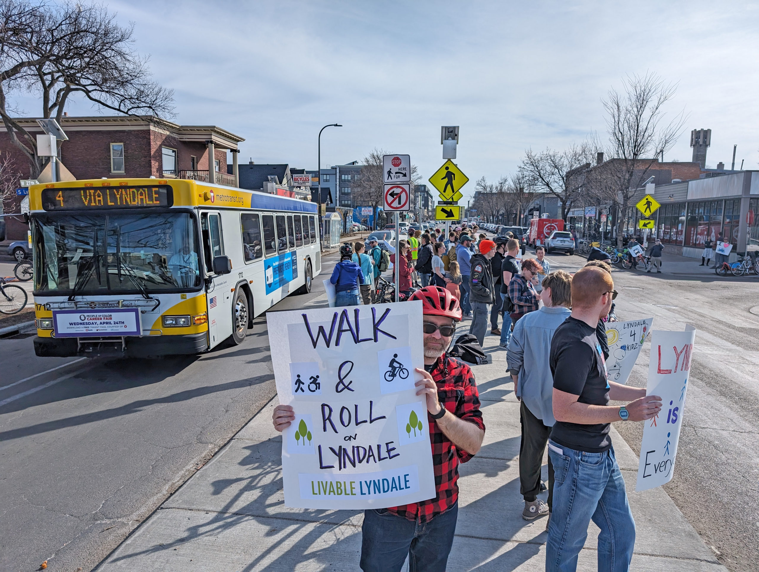 Community members rally in the median at Lyndale and 27th in Minneapolis as a Metro Transit bus drives by. A person holds a sign to support better walking and rolling on Lyndale. 