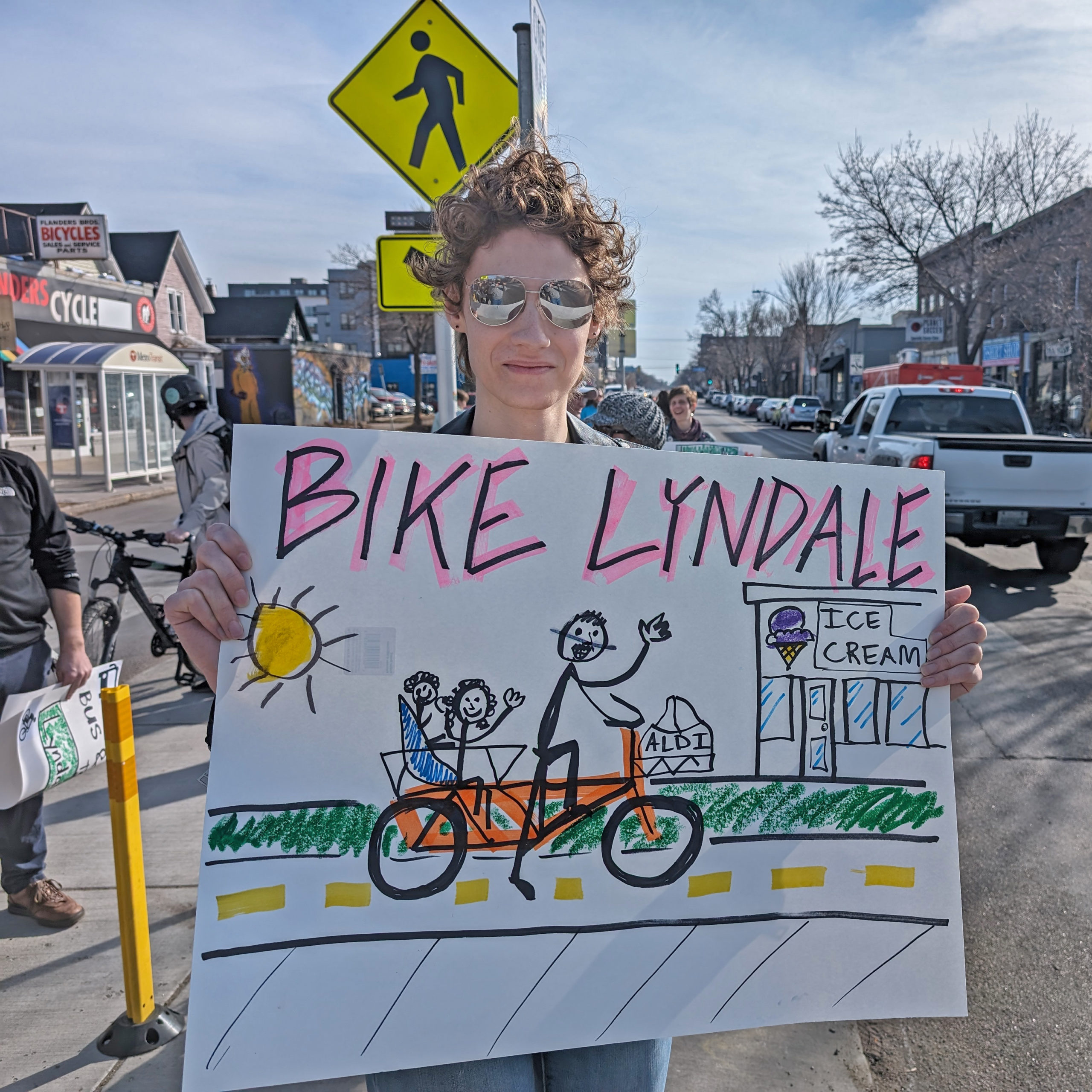 A community member at the Livable Lyndale rally in Minneapolis holds a hand-drawn sign that says Bike Lyndale. The sign features a drawing of an adult and two kids riding a cargo bike together on a separated bike lane and ice cream shop in the distance.