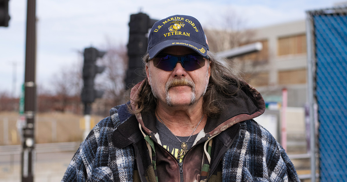 Dino stands outside in Saint Paul, MN, wearing sunglasses and a Marine Corps Veterans hat, about to transfer from a Metro Transit bus to a light rail train. The light rail station is visible in the background.