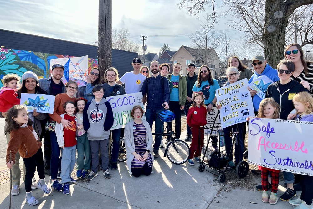 A multi-generational group pose for a photo at the corner of 27th and Lyndale Avenue, holding signs with slogans like "Lyndale is for people, bikes and buses" and Safe, Accessible Sustainable Lyndale"
