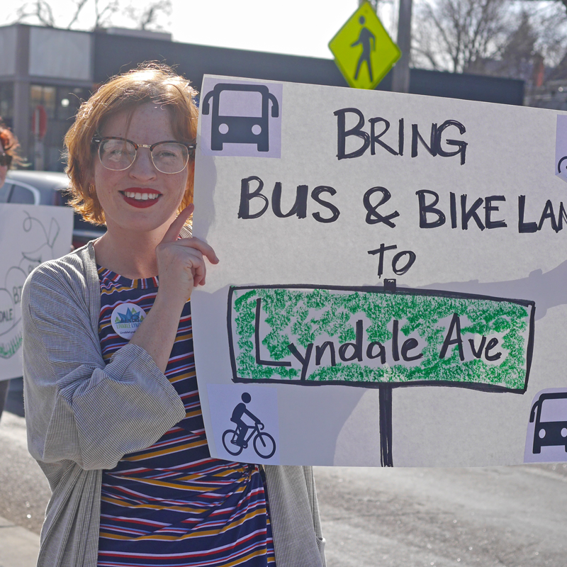 Stina Neel, a white woman wearing glasses and bright red lipstick, holds up a sign that says "Bring bus and bike lanes to Lyndale Ave"