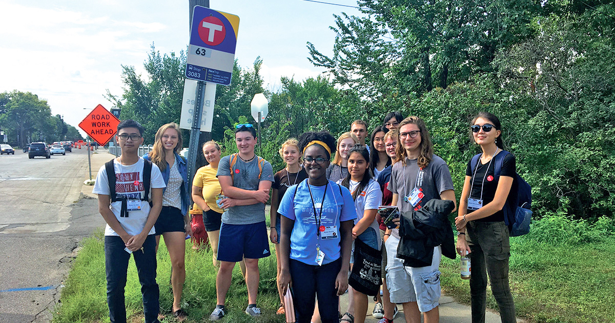 A group of fourteen young adults stands together at at Metro Transit bus stop. 