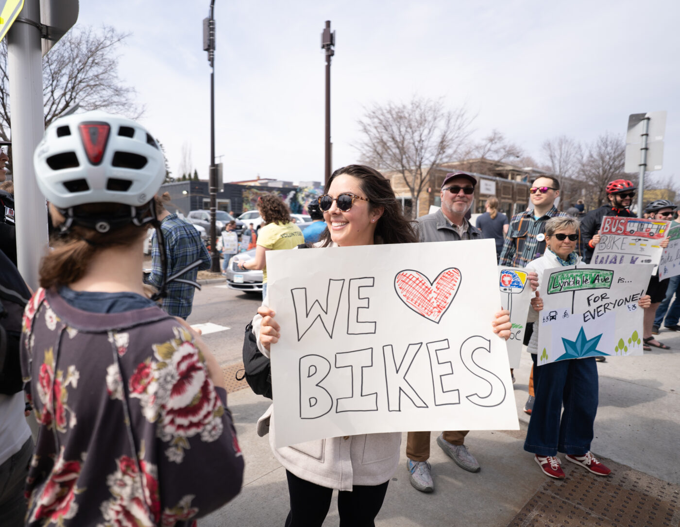 A smiling women with long dark hair and wearing sunglasses holds a sign that says "We [heart] Bikes."