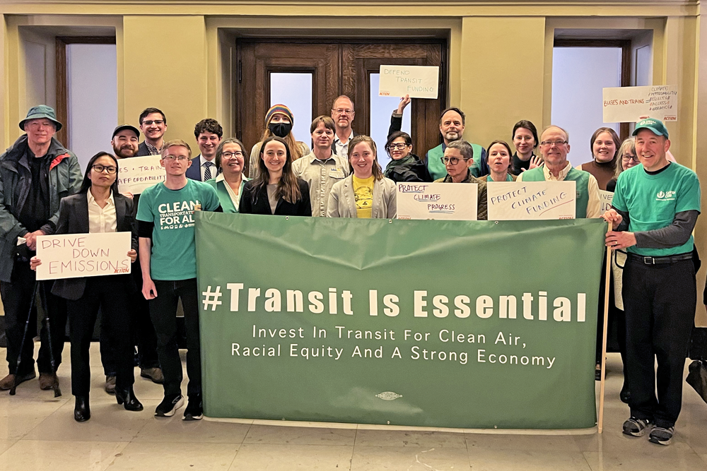 Advocates outside of a hearing room at the State Capitol holding a large green banner that says "Transit is Essential"