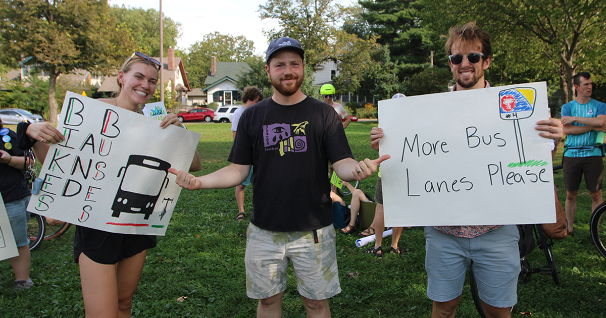 Three people pose at a rally, holding pro-bike lane and pro-bus lane signs