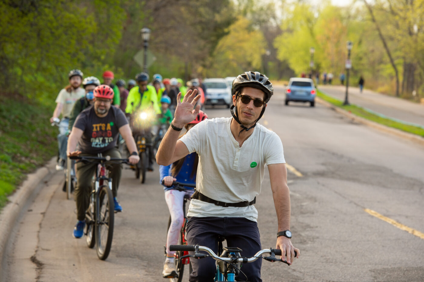 A person riding their bike waves at the camera with a group of other bikers behind them.